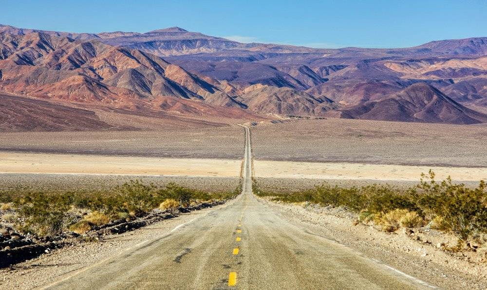 A straight, seemingly endless road stretches through the vast and arid landscape of Death Valley National Park, with rugged mountains in the distance under a bright, clear sky.