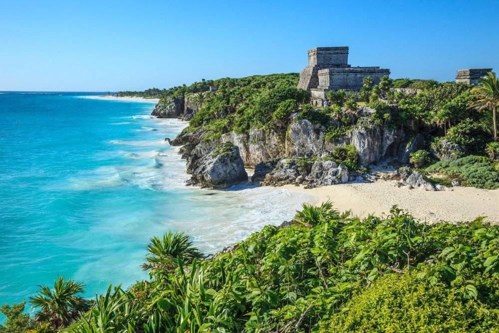 A scenic view from the ancient Mayan ruins in Tulum, overlooking a pristine white-sand beach and the turquoise waters of the Caribbean Sea.